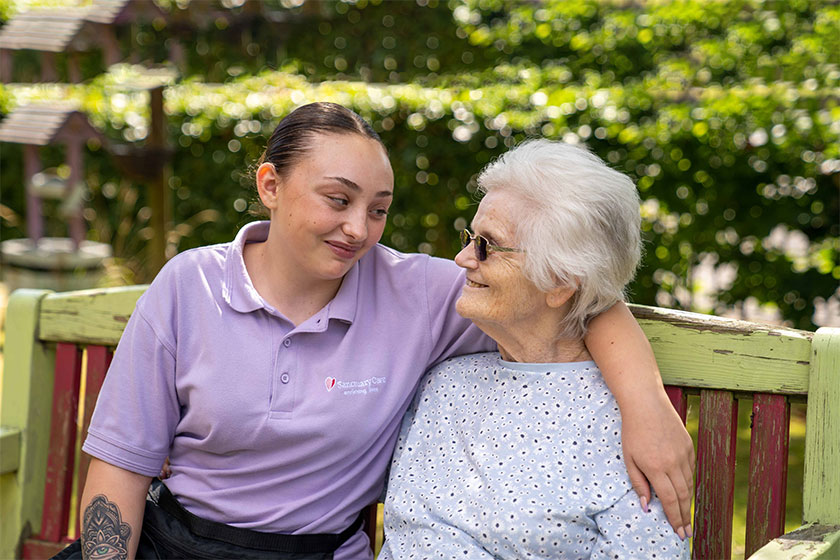 A female sanctuary care employee wearing a mauve branded t-shirt sitting on a bench with her arm around an elderly resident