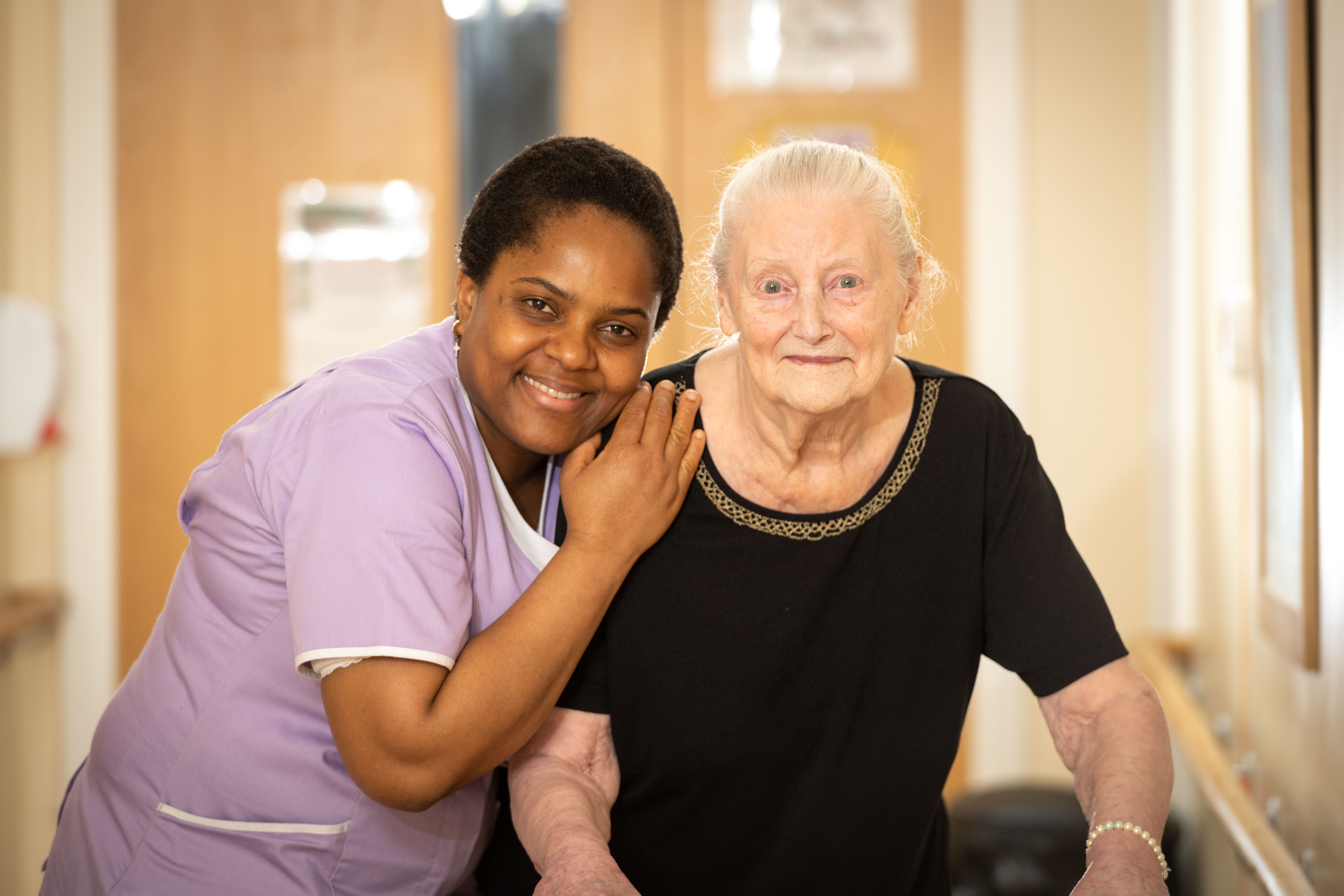 Carer and care home resident stand smiling in corridor 