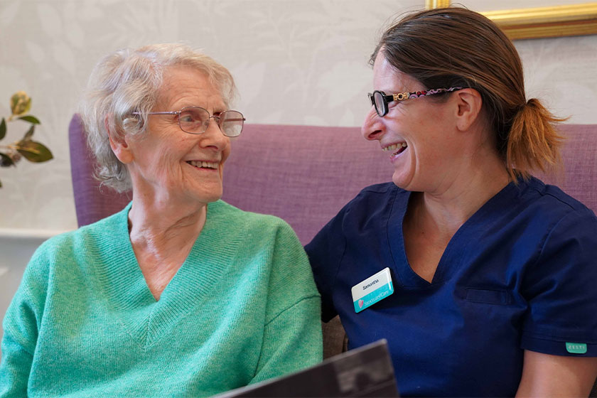 A female Sanctuary Care carer sitting on a purple sofa with an elderly resident. Both women are looking at each other and smiling.