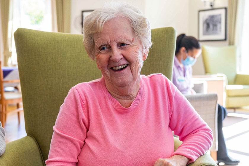 An elderly woman wearing a pink top sitting in a green armchair smiling. A care worker wearing a face mask can be seen in the background