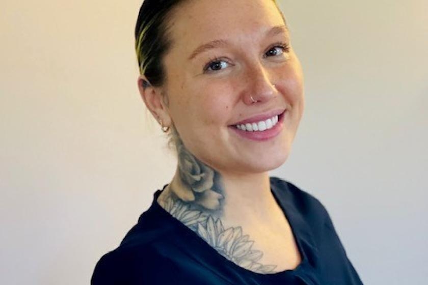 A female with their hair tied back with a tattoo on their neck smiling brightly into the camera