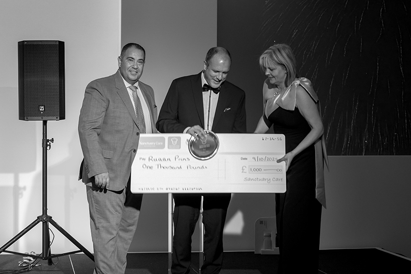 Chef Ruaan Prins wins award at Annual Chefs' Conference 2025 A black and white image of three individuals at an event. In the center, a man in a suit holds a large novelty check that reads "Mr. Riaan Puns, One Thousand Pounds." To his left, a man in a gray suit smiles, while to his right, a woman in an elegant dress looks on. The background features a softly lit setting, suggesting a formal occasion.