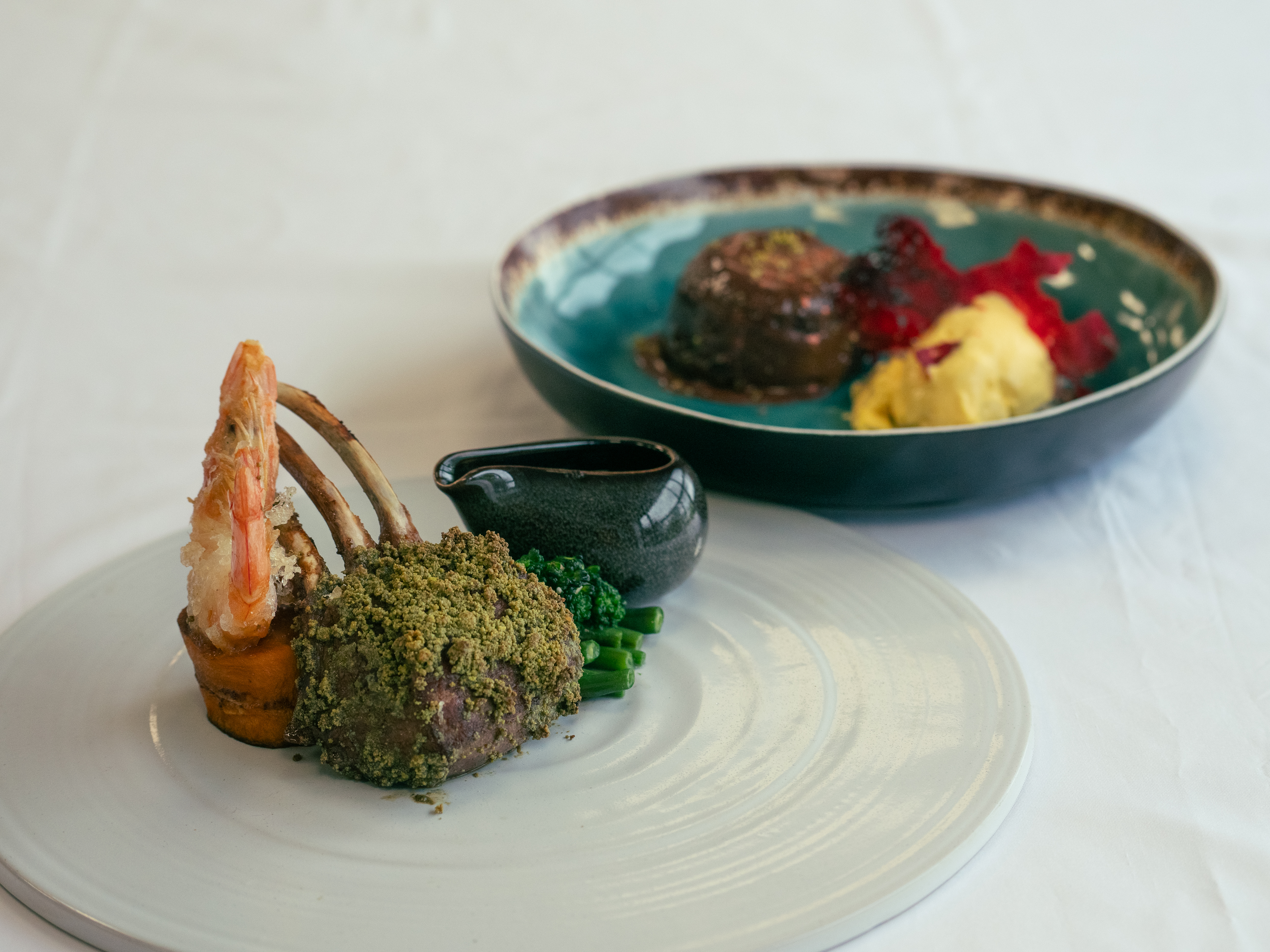 A beautifully plated gourmet dish featuring a crusted piece of meat, garnished with a large shrimp and accompanied by a small black sauce jug. In the background, a colorful plate showcases two portions of dessert, one with a creamy yellow texture and the other with a rich dark appearance, alongside vibrant red and pink garnishes.