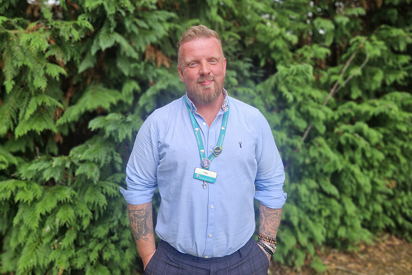 A man with a beard stands in front of a dense green hedge. He is wearing a light blue button-up shirt and has tattoos visible on his arms. A lanyard with a badge hangs around his neck. The background features lush foliage, creating a natural setting.