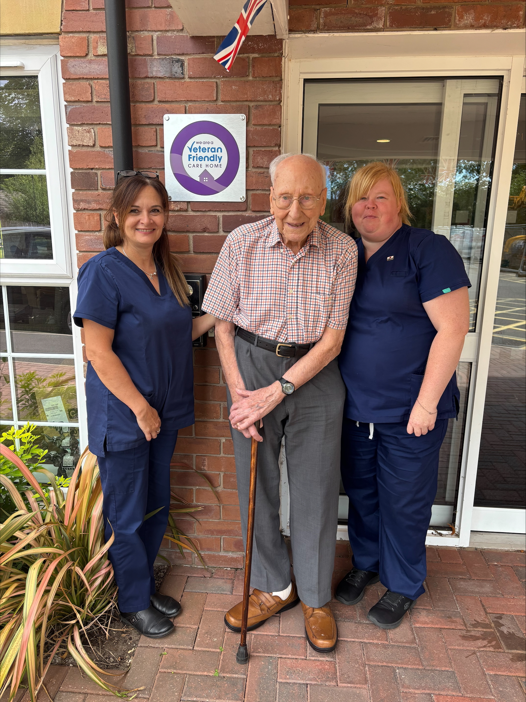 An elderly man stands in the center, holding a cane, and smiling. He is flanked by two female caregivers, both wearing navy blue scrubs.