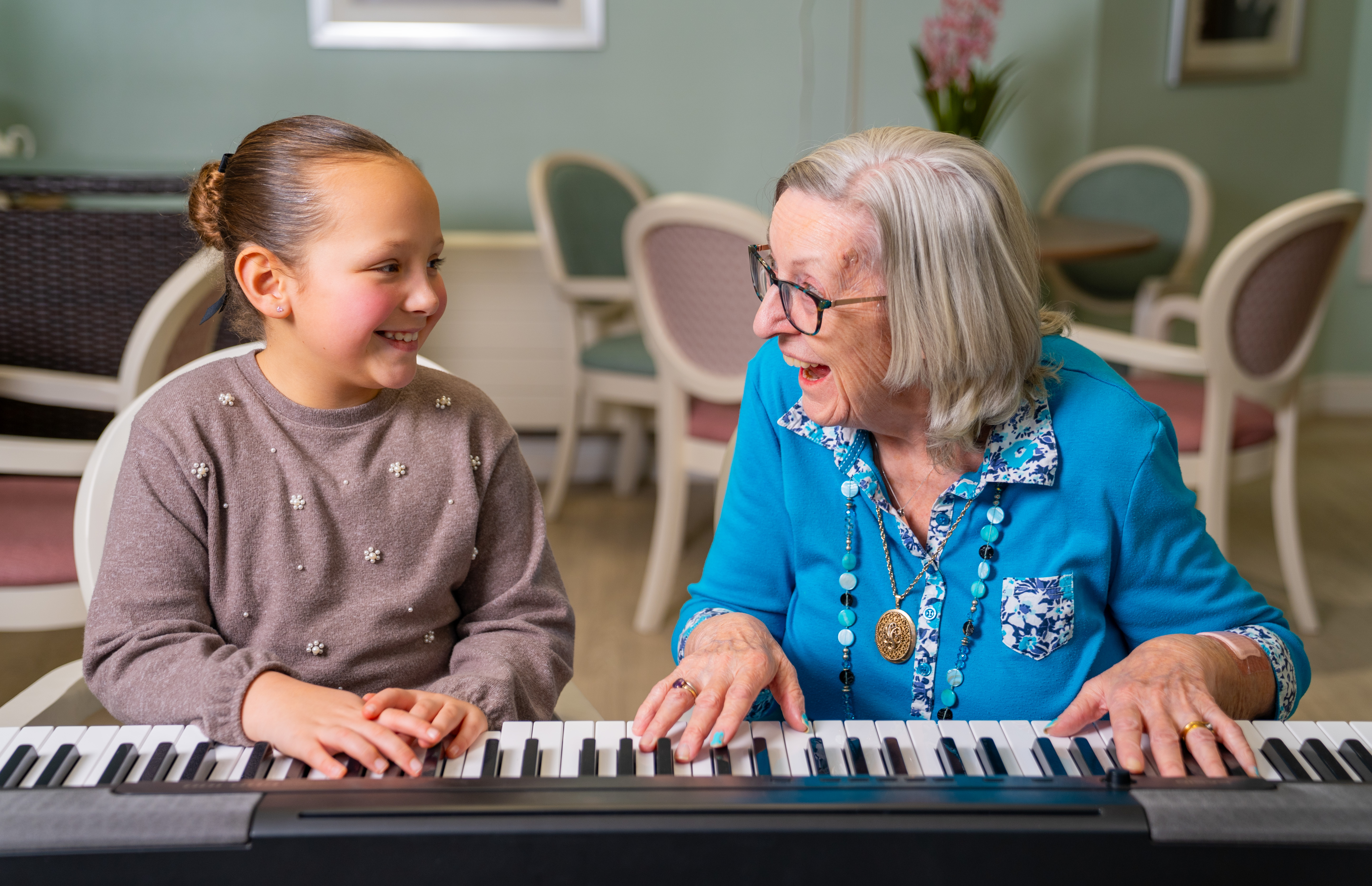 Young girl and resident Margaret smiling together while playing a keyboard piano in a cosy, pastel-coloured room