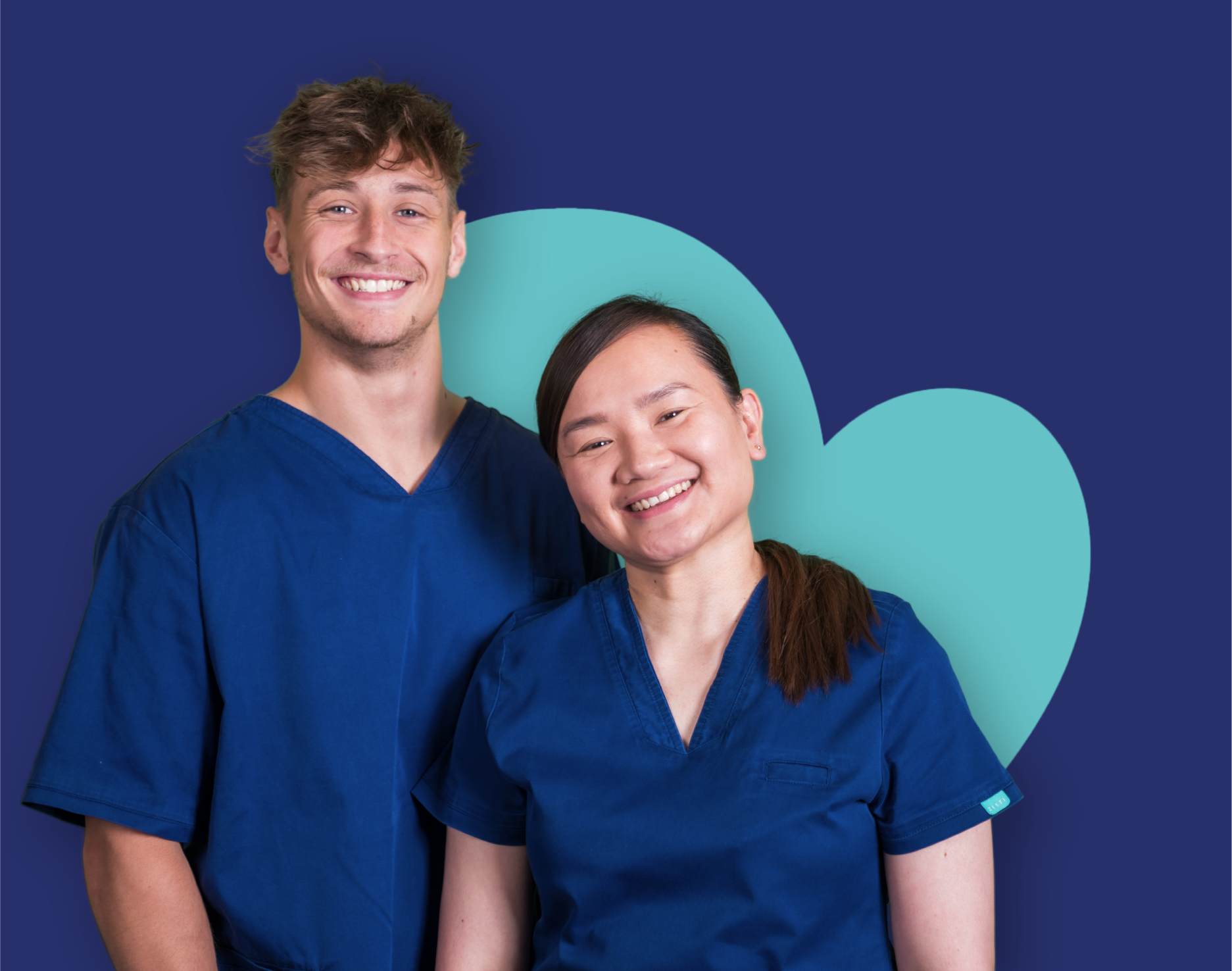 Two people in blue scrubs standing together and smiling in front of a large heart shape on a dark blue background.