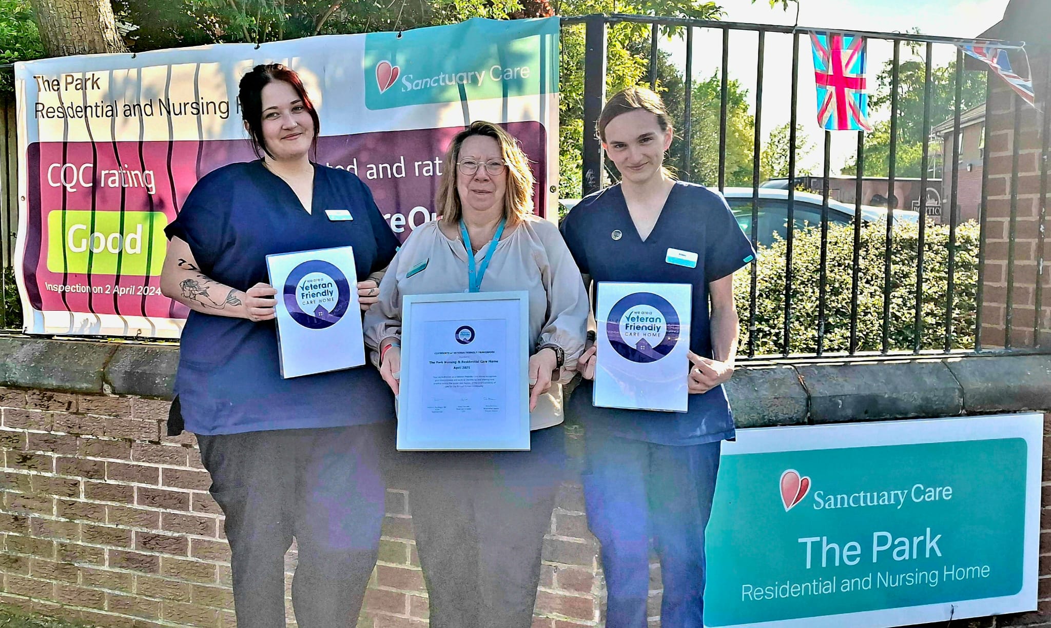 Three individuals stand in front of a brick wall and metal fence, holding certificates. Behind them is a banner that reads 'The Park Residential and Nursing Home' with a CQC rating of 'Good' as of 21 April 2023. Another sign reads 'Sanctuary Care The Park Residential and Nursing Home.' An English flag is visible in the background.