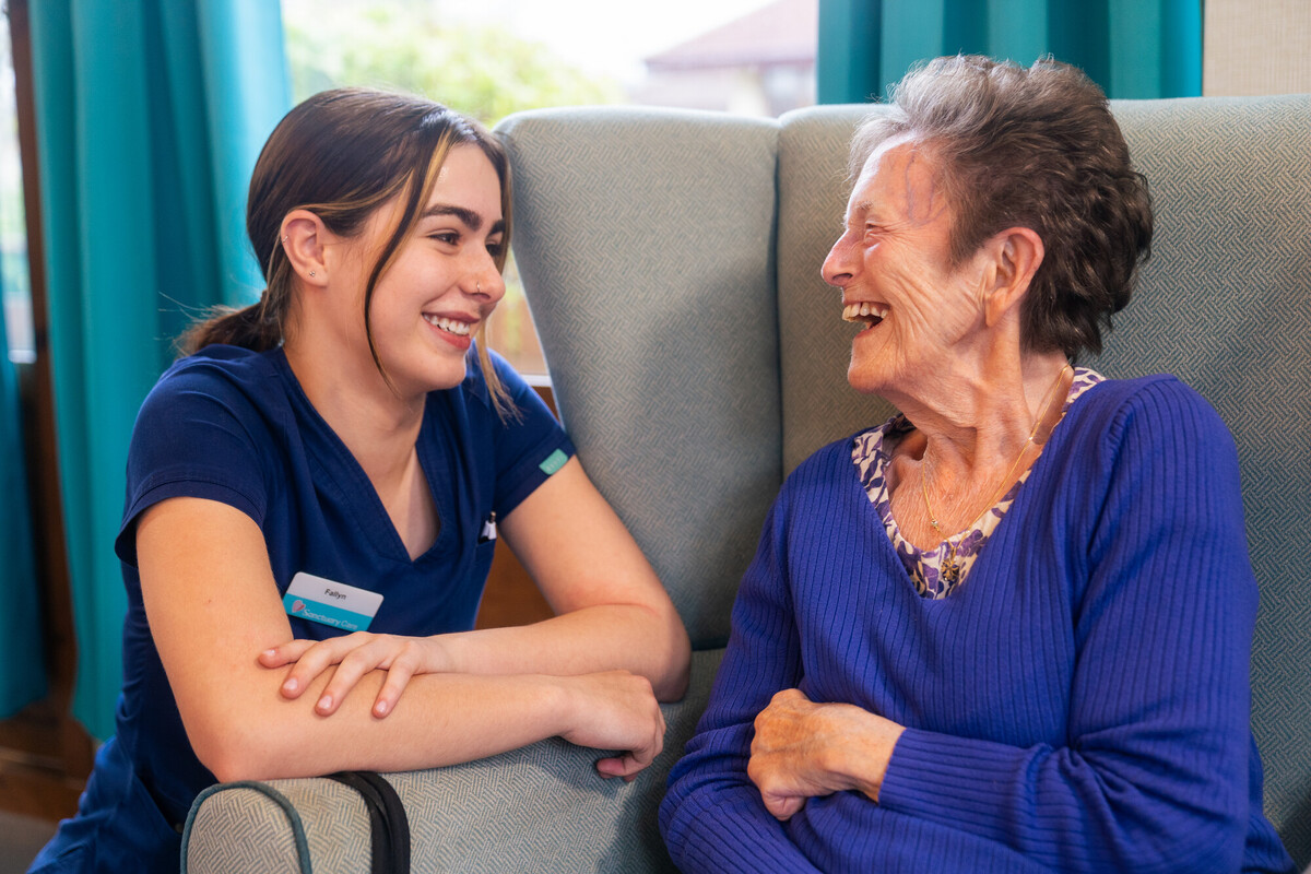 Care team member sitting with an older resident, talking together in a care home lounge