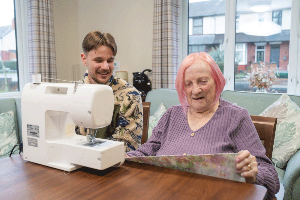 A person sitting at a table using a sewing machine, with another person seated beside them offering support. The table holds fabric being guided through the machine, and the scene takes place in a bright room with large windows and patterned curtains.