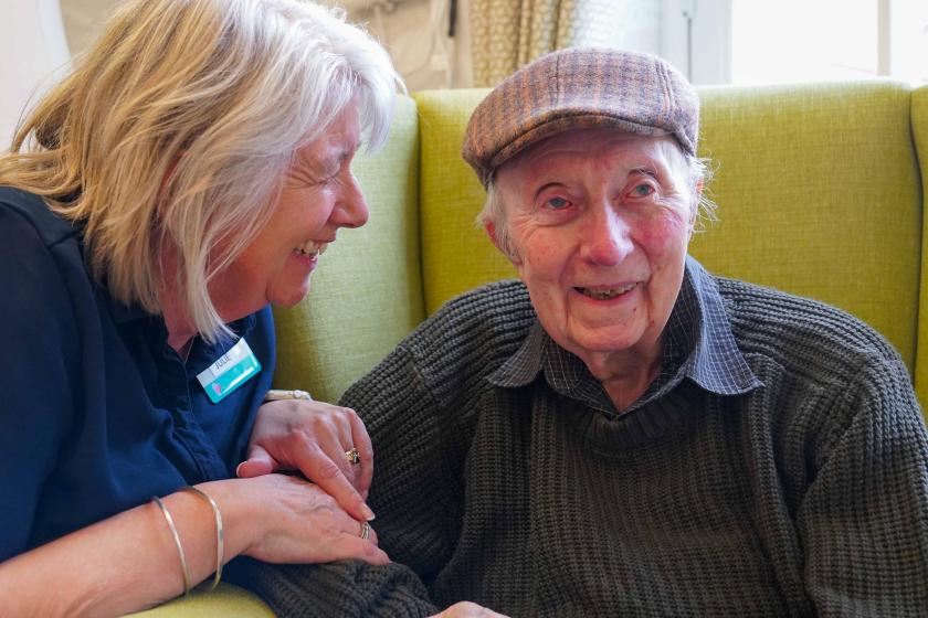 A male resident sat in a comfortable yellow arm chair with a member of staff smiling with him and comforting him