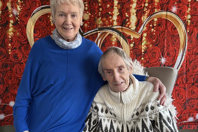 Sanctuary resident Tom sat in an armchair with his wife Clare sat on the arm of the chair in front of a backdrop of roses.