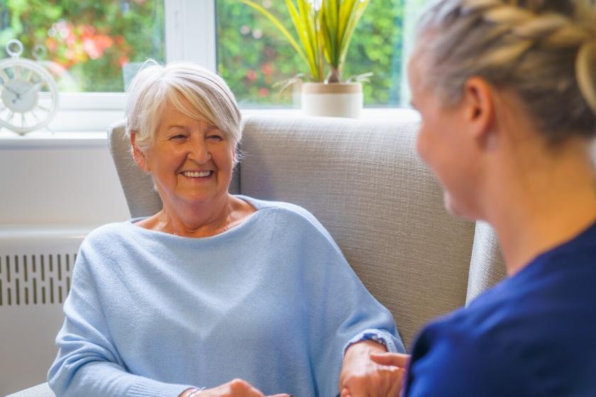 Resident Yvonne wearing a pale blue jumper sat angled in a large arm chair smiling at staff member Leanne