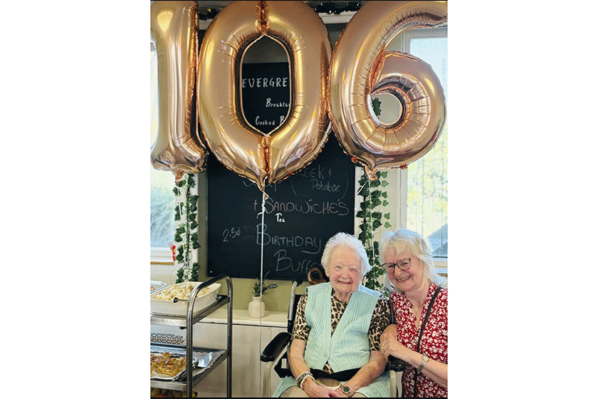 Maria Blagden and Resident Joyce Penfold sitting at The Green Residential Care Home in Redruth Cornwall. They are sat together and smiling with 106th birthday balloons behind them. They are both smiling