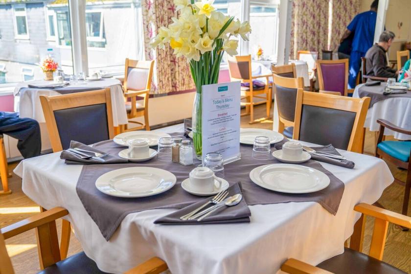 A bright, welcoming dining area at The Woodland, with neatly set tables, fresh flowers, and large windows letting in natural light. Creating a warm, friendly, and community-focused atmosphere.