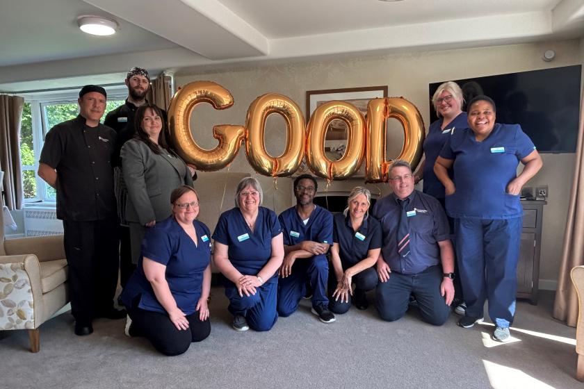 A group of people pose indoors with gold balloons spelling "GOOD"; some are kneeling, others standing, in a bright room with a window and TV.