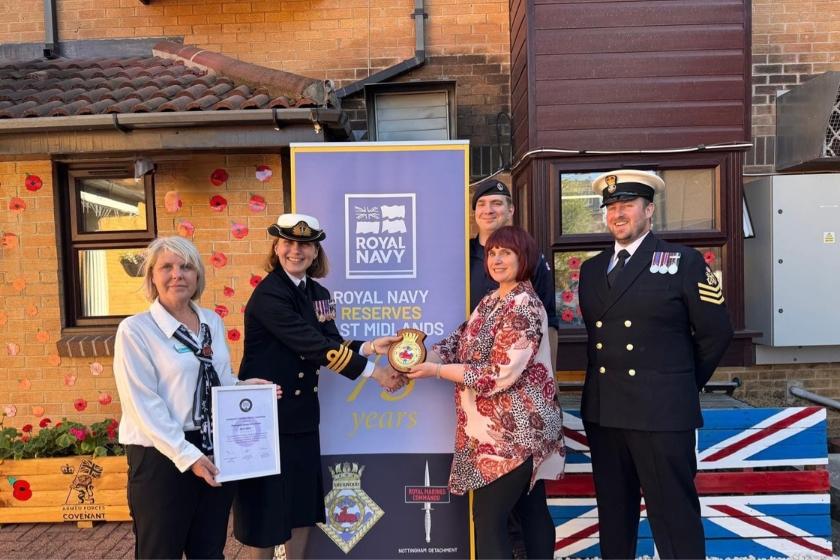 Four individuals standing in front of a banner reading 'ROYAL NAVY RESERVES WEST MIDLANDS.' Two are in Royal Navy uniforms; one holds a certificate, and another hands an object to a person in civilian clothing. A brick building with poppy decorations and a Union Jack flag design is in the background.