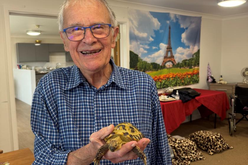A resident of Regent care home is holding a small tortoise in one hand. The person is wearing a blue checkered shirt and standing in a room with beige carpet, red tablecloths, and leopard print cushions on the floor. A large poster of the Eiffel Tower with tulips in the foreground hangs on the wall behind them.