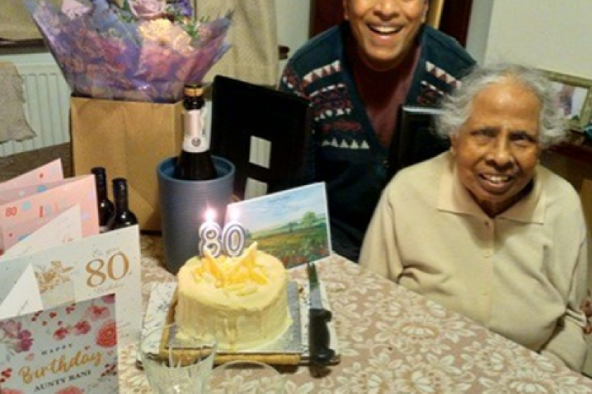 An older person and another individual seated at a table decorated for an 80th birthday celebration, featuring a cake with '80' candles, birthday cards, flowers, and a bottle of champagne.