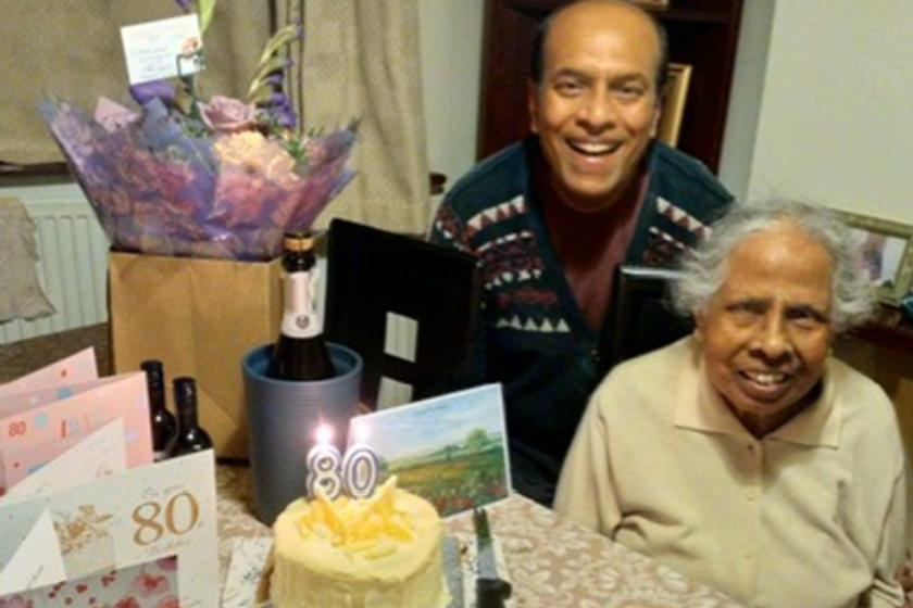 An older person and another individual seated at a table decorated for an 80th birthday celebration, featuring a cake with '80' candles, birthday cards, flowers, and a bottle of champagne.