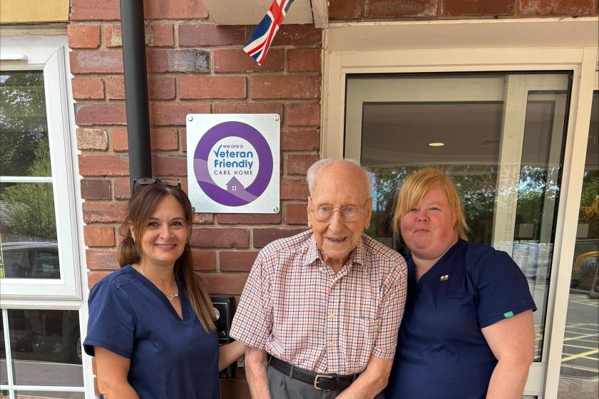 Three people standing outside a building marked 'Veteran Friendly Care Home.' The central person holds a cane, flanked by two in blue uniforms. A British flag is above a purple-circled sign.