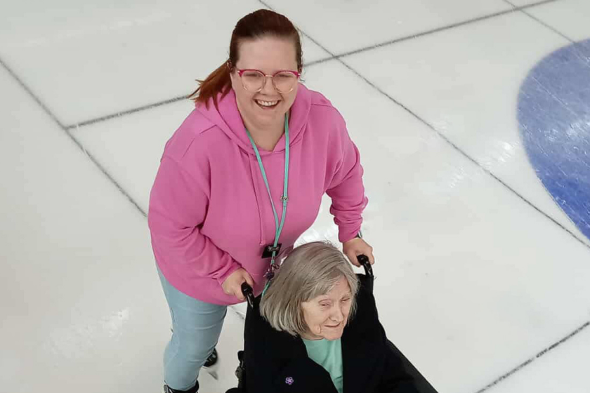 Activities coordinator Kirsty smiles at the camera as she pushes a resident around the ice rink in her wheelchair