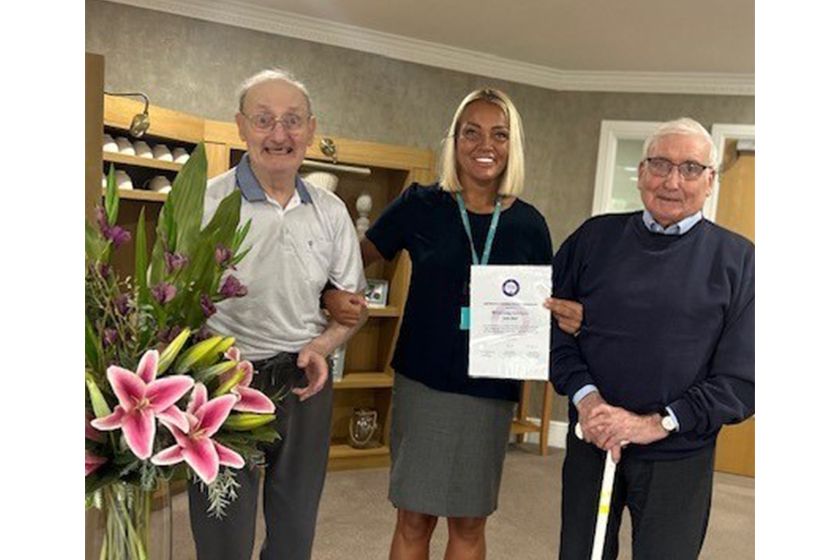 Three people stand indoors, with the person in the middle holding a certificate. A bouquet of flowers sits on a small table to the left. Shelves and a door are visible in the background.