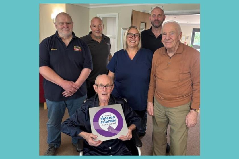 Residents and staff at a Care Home with a resident sat in the front of the frame holding a veteran friendly care home poster