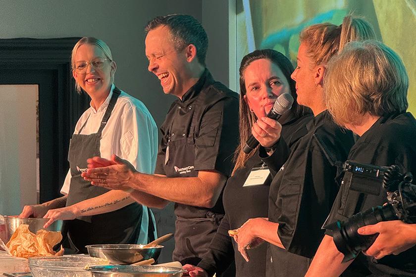 Chefs on stage doing live cooking demonstration for attendees at the Chefs Conference A group of five chefs stands behind a kitchen counter, engaged in a lively cooking demonstration. They are wearing black and white chef uniforms and smiling as they interact with each other. The background features a large screen displaying a food-related image. The atmosphere appears energetic and collaborative, with various cooking utensils and ingredients visible on the counter.
