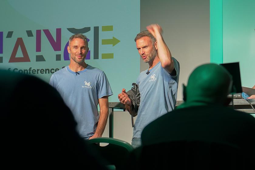 Two men in light blue t-shirts are standing on a stage, engaging with an audience during a conference. One man is gesturing with his hand, while the other appears to be listening. In the background, there is a colorful logo or banner that reads "CHANGE" along with the words "Conference." The audience is partially visible in the foreground, with their backs to the camera.