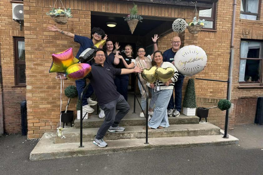 A group of seven people is gathered on the steps of a brick building, celebrating with balloons. They are smiling and posing playfully,