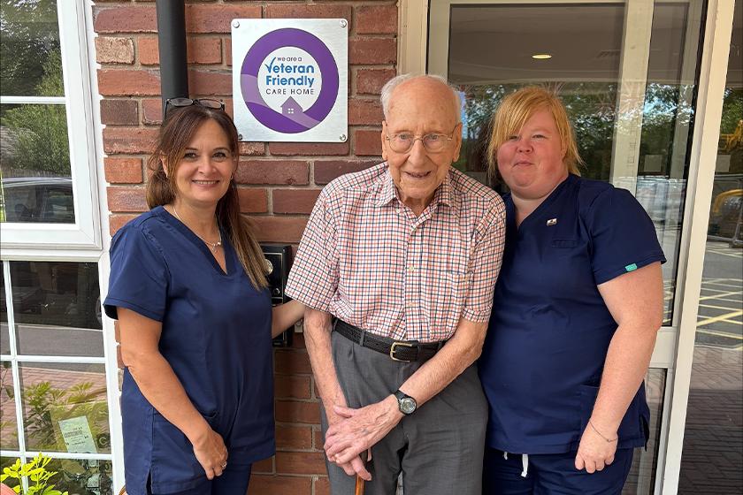 An elderly man stands in the center, holding a cane, and smiling. He is flanked by two female caregivers, both wearing navy blue scrubs.