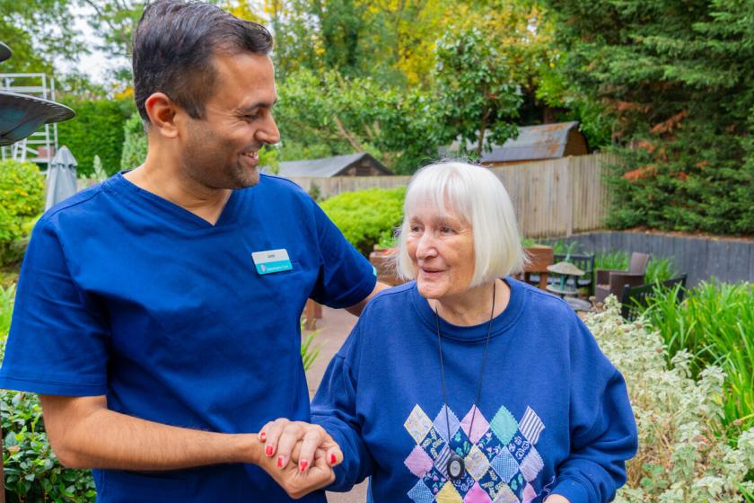 Carer-and-Resident-Haven-Care-Home A healthcare worker in a blue uniform smiles and holds hands with an elderly woman in a blue sweater adorned with a diamond pattern. They are standing in a lush garden with greenery and outdoor seating in the background. The scene conveys warmth and companionship.