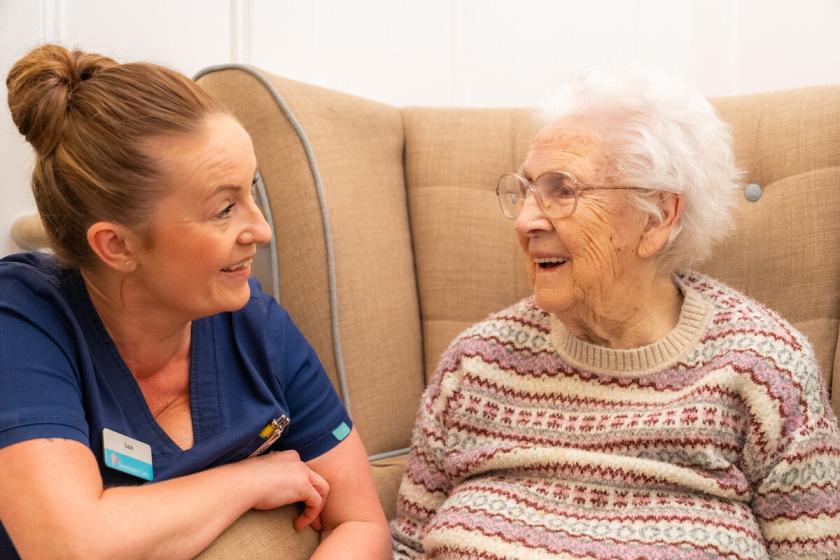 Care worker sitting beside an older adult on a sofa, both facing each other in conversation.