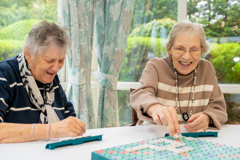 Residents-Enjoying-Scrabble Two elderly women are sitting at a table, enjoying a game of Scrabble. They are both smiling and engaged in the game, with one woman placing a tile on the board while the other laughs. The background features large windows with greenery visible outside, creating a bright and cheerful atmosphere. The women are dressed casually, with one wearing a striped scarf and the other a beige sweater with a necklace.