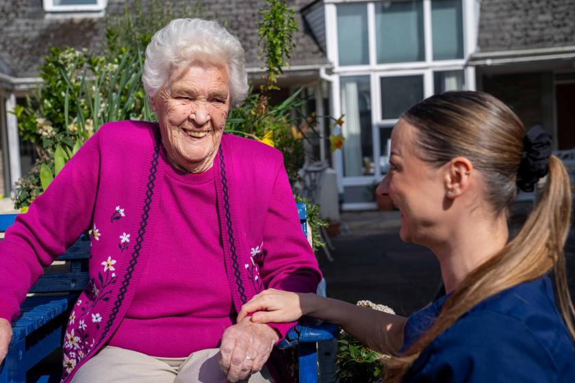 An woman with white hair is sitting on a blue bench, wearing a bright pink cardigan adorned with floral patterns. She is smiling warmly at a caregiver, who is seated beside her, holding her hand. The caregiver has long brown hair tied back and is wearing a dark blue uniform. In the background, there are plants and a building with large windows, suggesting a pleasant outdoor setting.