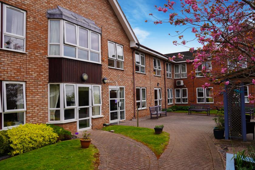 A brick building with multiple windows and a welcoming entrance is surrounded by a landscaped garden. The pathway is lined with green grass and flower beds, and a flowering tree adds a pop of color. Benches are placed along the path, creating a serene outdoor space.