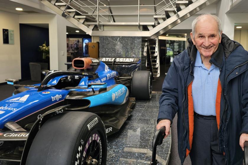 An elderly man stands next to a vibrant blue racing car, looking pleased and happy.
