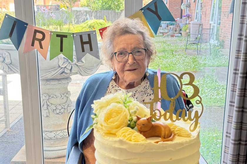 An elderly woman smiles while sitting in front of a birthday cake 