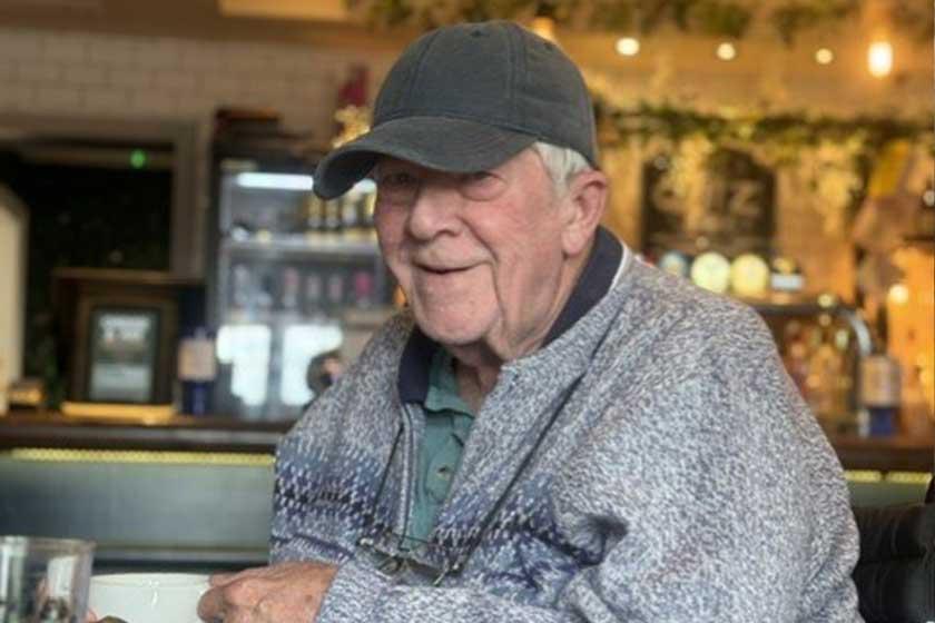 An elderly man seated at a table, holding a cup of coffee and smiling softly.