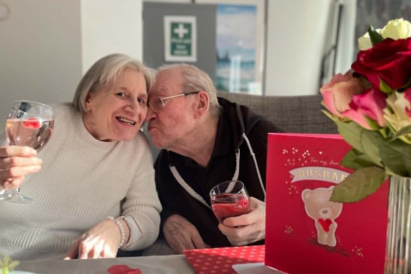 An elderly couple is sitting closely together at a table, smiling and enjoying drinks. The woman is holding a glass and leaning towards the man, who is giving her a kiss on the cheek. The table is decorated with a bouquet of flowers, cupcakes, and a large greeting card featuring a teddy bear and the word "Valentine." A red balloon is visible in the background, adding to the festive atmosphere.