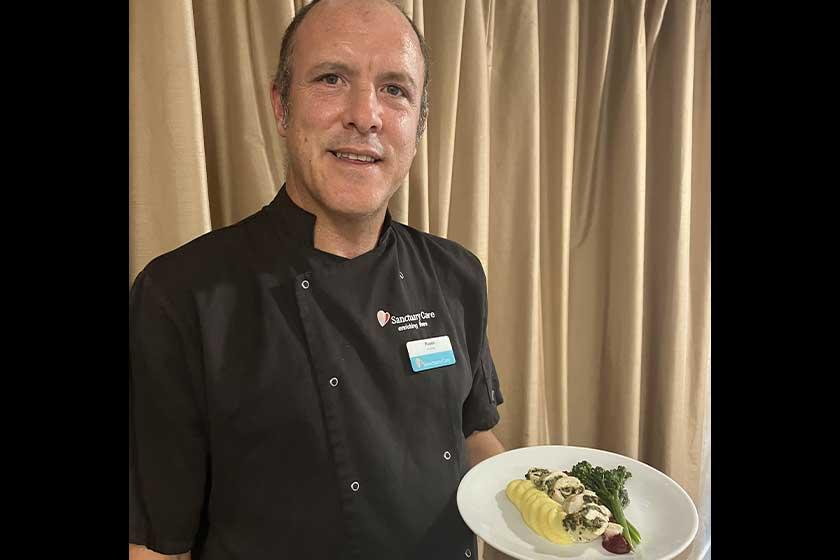 A chef wearing a black uniform with the "Sanctuary Care" logo is presenting a plate of food.
