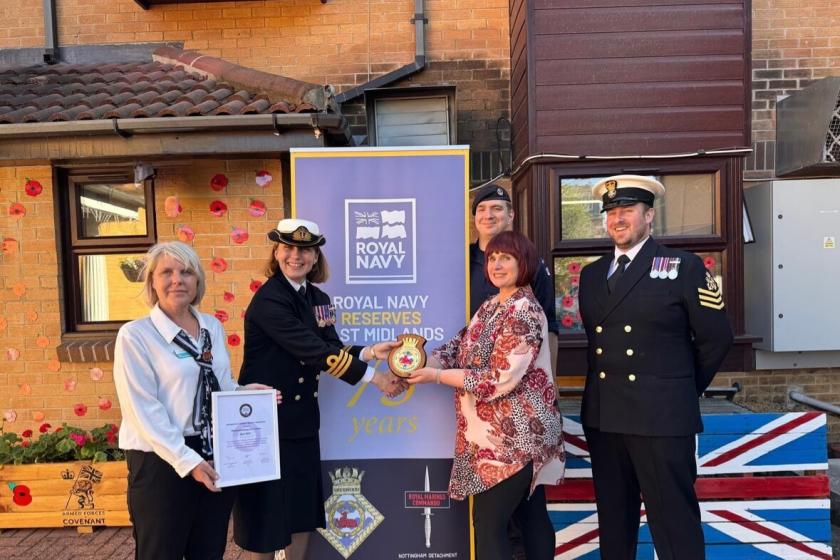 A group of five individuals stands in front of a banner celebrating 75 years of the Royal Navy Reserves in the West Midlands. The banner features the Royal Navy logo and is decorated with a Union Jack motif. Two members of the group are in naval uniforms, while the others are dressed in civilian attire. They are holding a commemorative plaque and a badge. The background includes a brick building and decorative elements, such as red paper poppies.