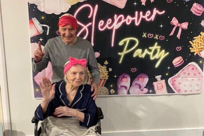 Jim and Ina Sleepover A person seated in a wheelchair and covered with a blanket poses in front of a pink-themed "Sleepover Party" backdrop, accompanied by another person standing behind. The display features pink balloons, neon-style lettering, and illustrations of sleepover items including slippers, bows, snacks, and beauty products.