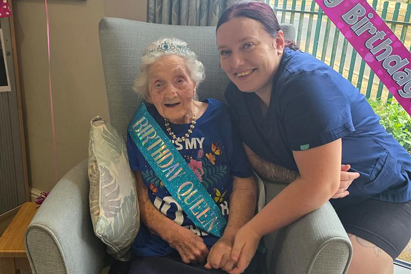 A smiling elderly woman wearing a "Birthday Queen" sash and a floral shirt sits in an armchair, with a young woman kneeling beside her, both smiling. A pink birthday balloon and several framed photos are visible in the background.
