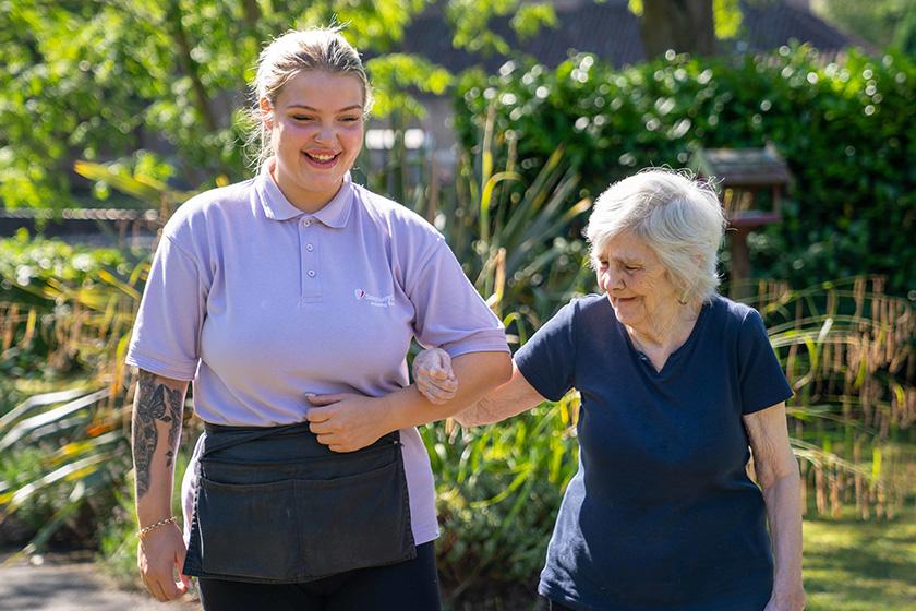 A care worker smiling while guiding an elderly lady through the gardens.