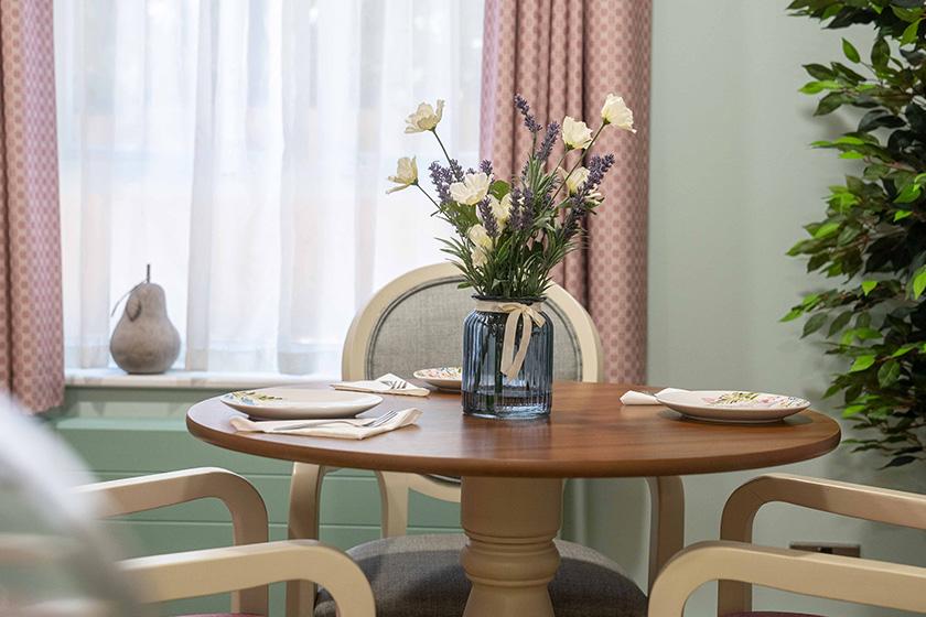 A peaceful seating area with numerous chairs located around the wooden circular dining table. It is decorated with fresh flowers to completement the pale colour pallet of the room.