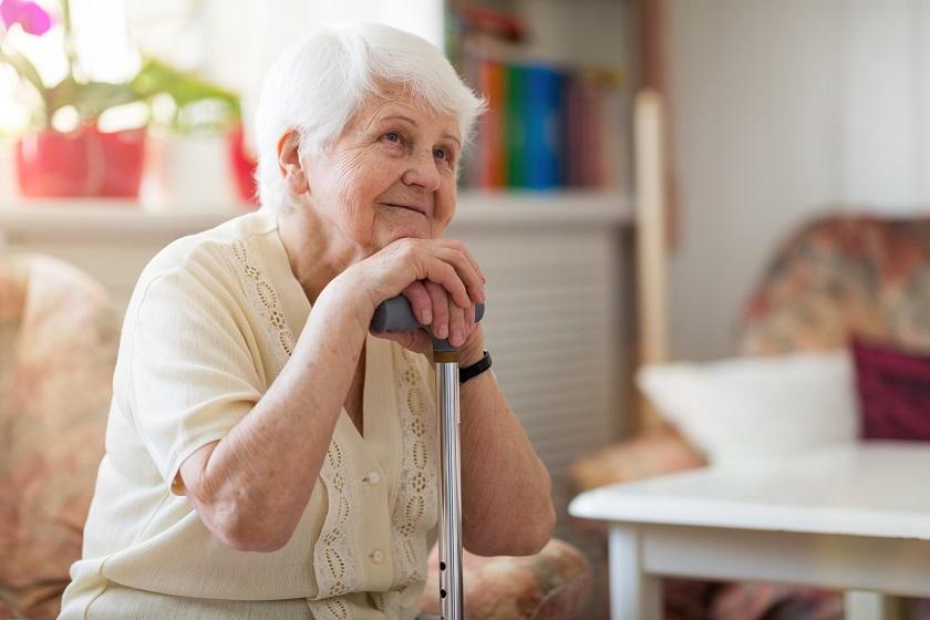 An elderly lady sits on a chair while leaning on her walking stick