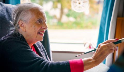 An elderly woman sitting on a sofa and smiling while painting with a paint brush