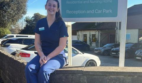 A young woman wearing blue medical scrubs sitting on a brick wall in front of a sign that reads 'Trevarna Residential and Nursing Home'